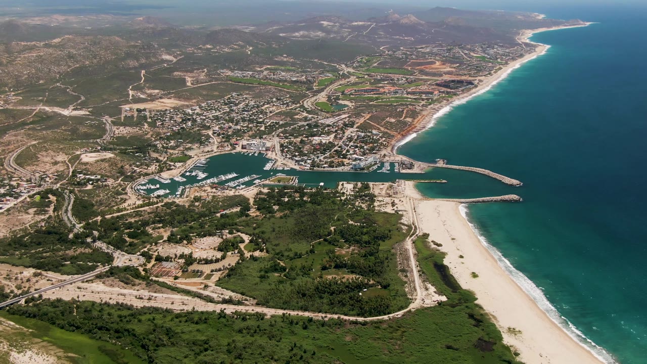 Aerial view of marina, sandy beach, turquoise ocean, town roads, and inland hills.