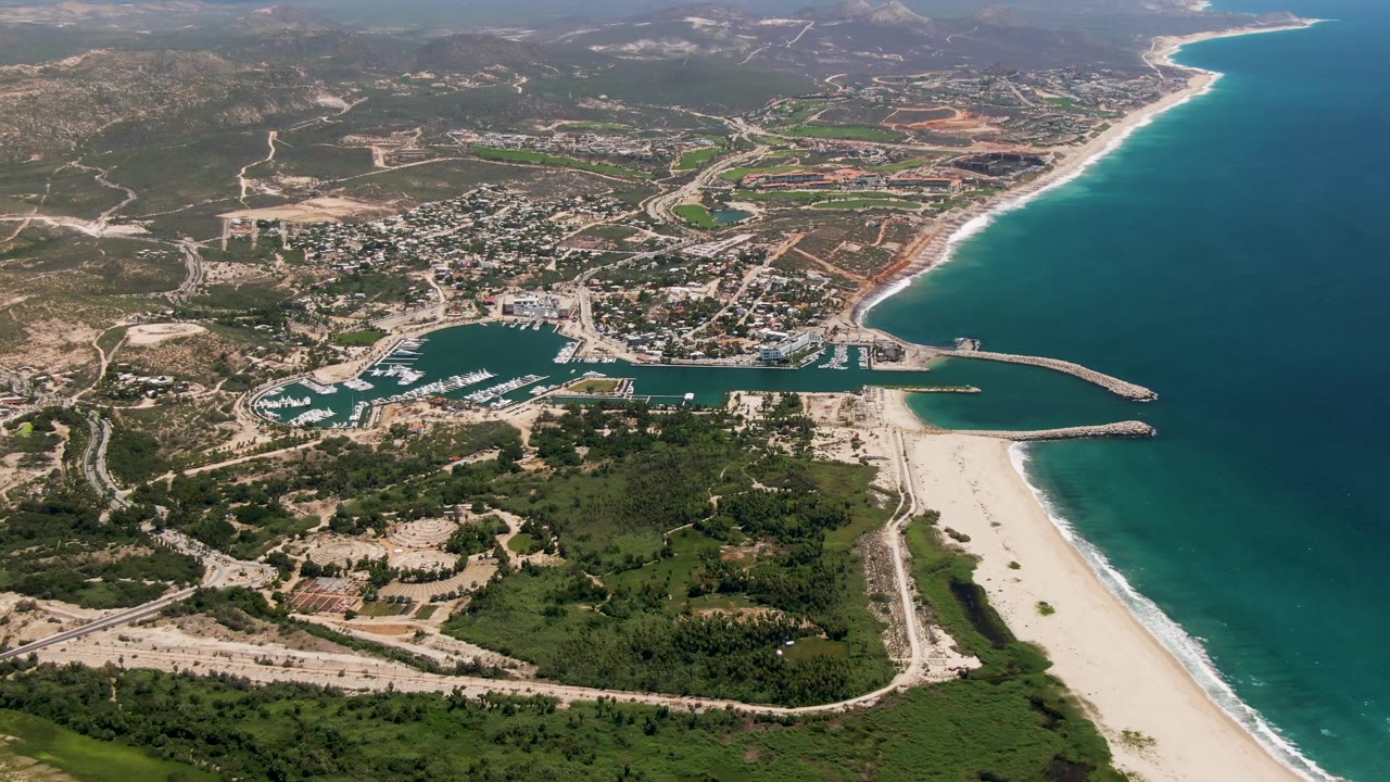 Aerial frame in San Jose Del Cabo with the marina slightly more centered, boats lined inside the basin, beach and surf along the right edge, roads and buildings inland, and no visible people.