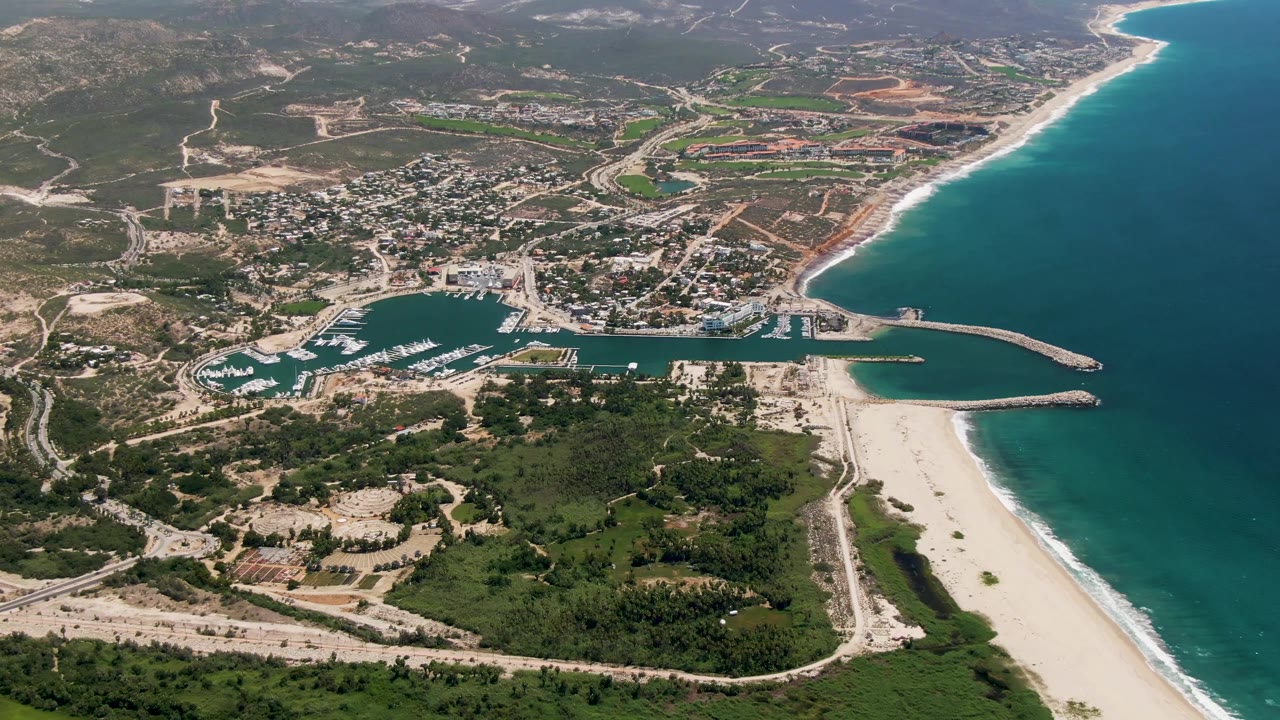 San Jose Del Cabo aerial frame with the marina entrance and breakwaters clear in the lower middle, the beach running up the right side, town blocks inland, dry hills beyond, and no visible people.