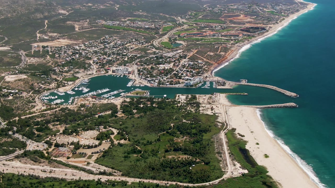 Mid-clip aerial frame over San Jose Del Cabo showing the marina and boat slips larger in view, pale beach and surf to the right, roads and low development inland, and no visible people.
