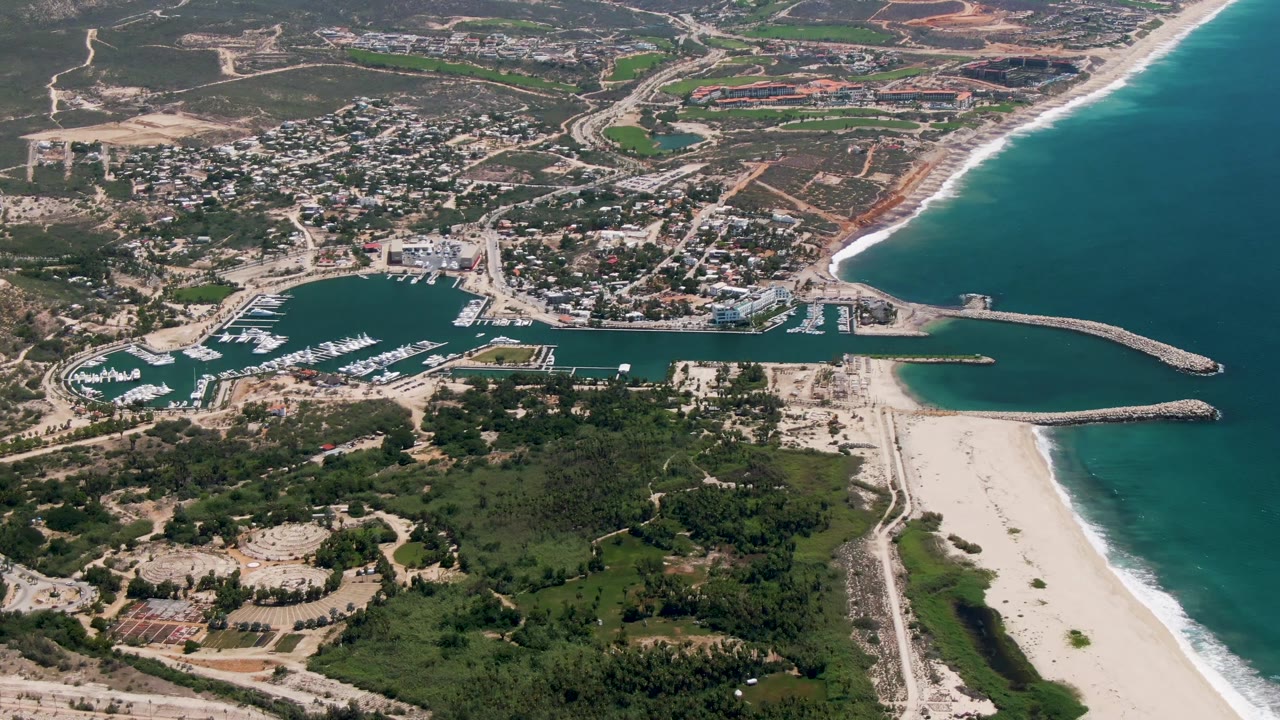 Opening aerial frame over San Jose del Cabo shows the marina basin, docked boats, curved breakwaters, sandy beach, green land, town roads, and no visible people.