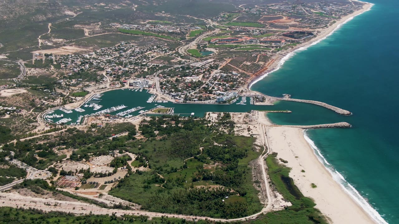 High aerial frame of San Jose Del Cabo with the marina centered, breakwaters reaching into turquoise water, a pale beach below, and no visible people.