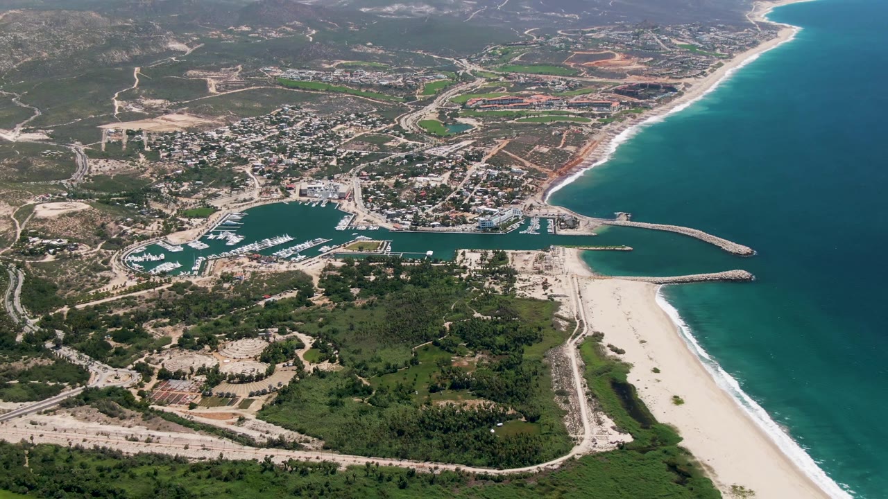 Aerial view over San Jose Del Cabo with the town and marina beside a long curving beach, open ocean on the right, green land below, and no visible people.
