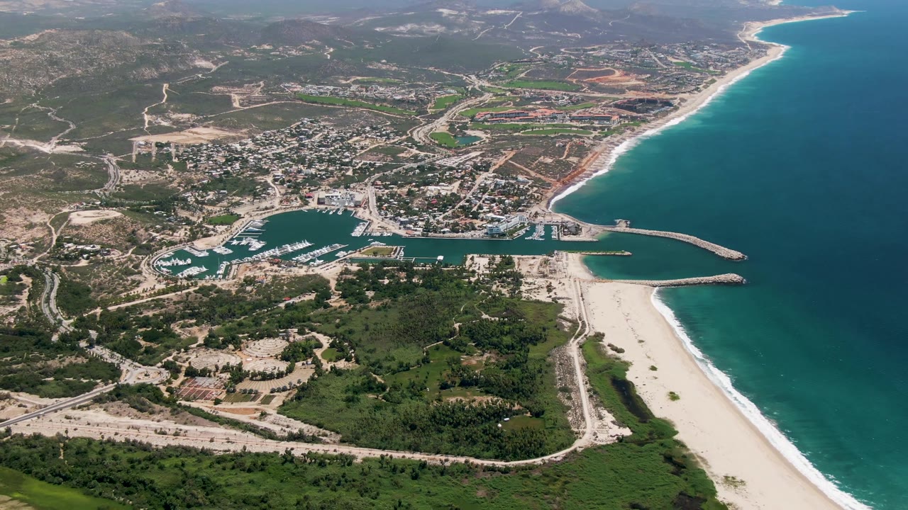 Wide San Jose Del Cabo aerial with more ocean visible to the right, the marina and beach in the lower center, roads across the inland terrain, and no visible people.