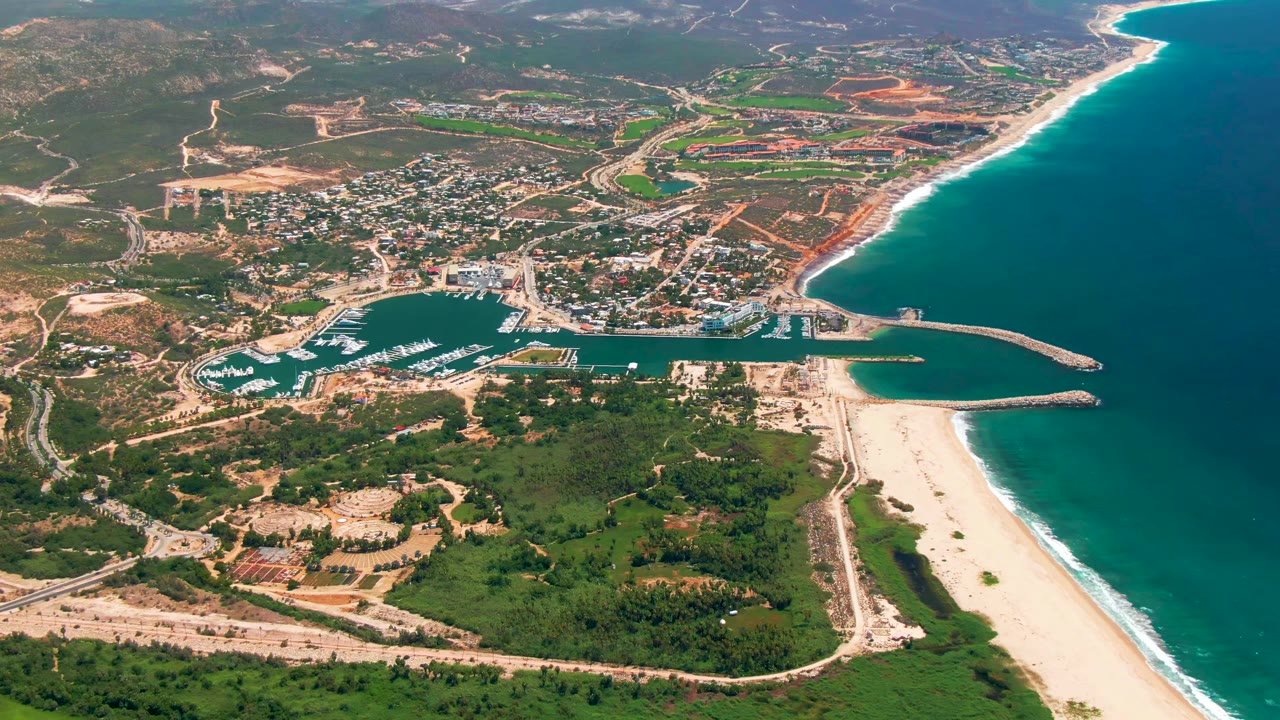 Opening aerial frame over San Jose Del Cabo, Mexico shows a marina packed with boats beside a long sandy beach, turquoise ocean, roads, greenery, inland buildings, and no visible people.