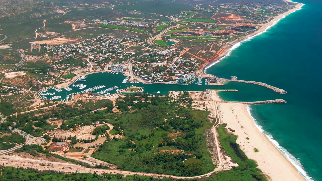 At 0:01, the San Jose Del Cabo aerial holds on the marina and breakwaters, with the sandy beach and white surf line along the right side and no visible people.