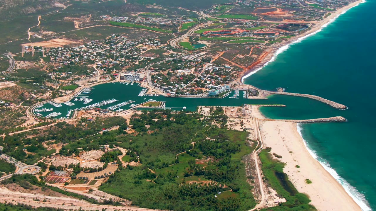 At 0:02, the frame shows the marina entrance between two rock arms, boats in slips, a bright beach strip, town roads, inland terrain, and no visible people in San Jose Del Cabo.