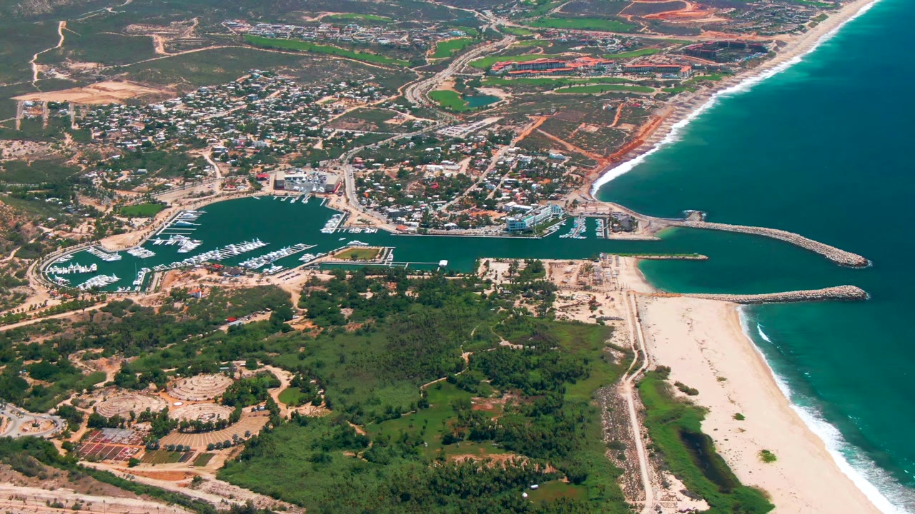 Aerial frame in San Jose del Cabo with white boats clustered in the marina, a bright sand beach along the right edge, surf on blue water, inland roads, and no visible people.