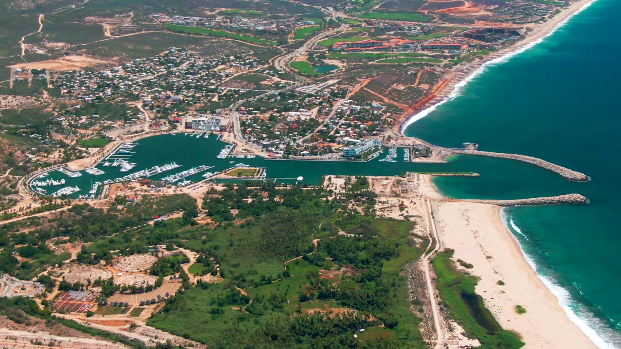 Aerial frame over San Jose del Cabo showing the harbor entrance and curved breakwaters, long sandy beach, turquoise-blue ocean, inland town streets, exposed construction soil, and no visible people.
