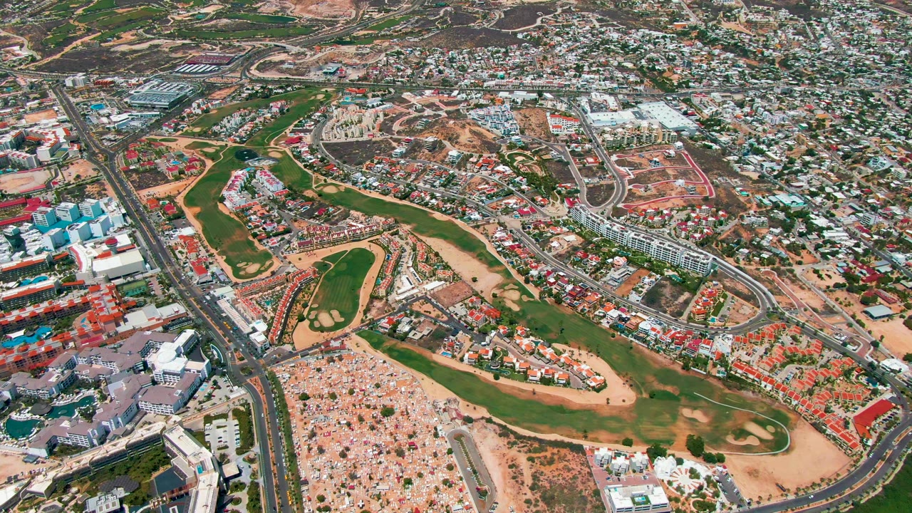Aerial frame over San Jose del Cabo showing green fairways through the center, paved roads, tan construction lots, dense buildings, distant hills, and no visible people.