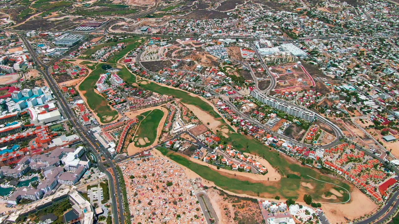San Jose del Cabo appears from above with fairway-shaped green areas, red-roof and light-roof buildings, a main road corridor, tan lots, and no visible people.