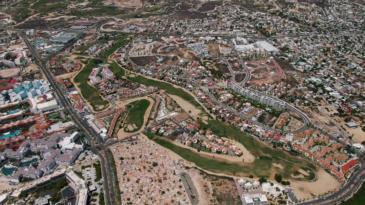 Opening aerial frame over San Jose Del Cabo showing a large crowd on sandy ground beside green fairways, roads, and dense low-rise buildings.