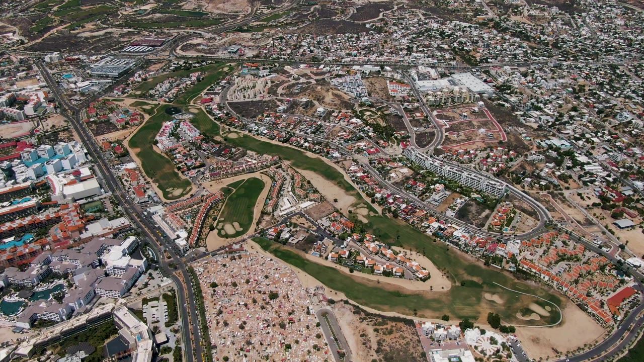 Wide drone view of fairways, roads, buildings, and a large gathering.
