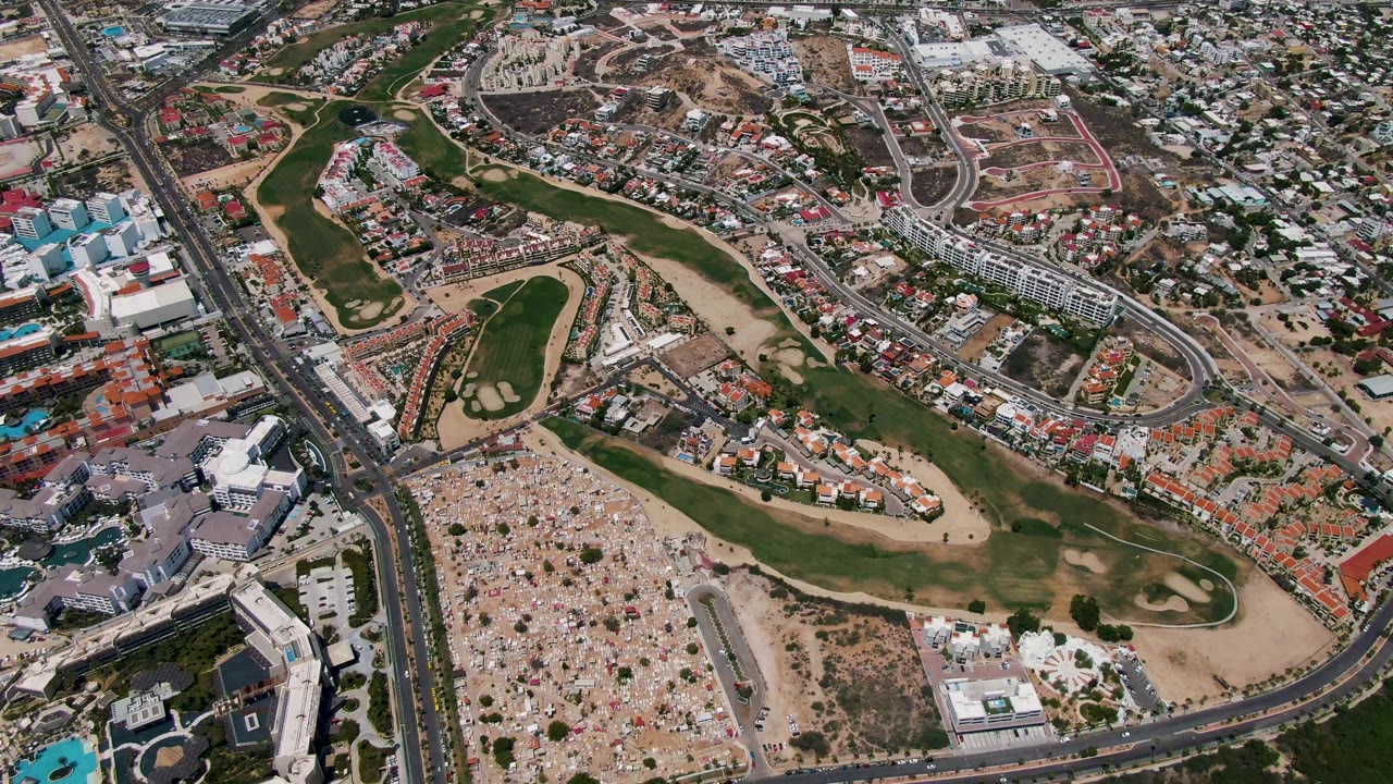 High aerial view in San Jose del Cabo showing a green golf course, sand traps, curving roads, residential blocks, commercial structures, dry lots, pools, and no visible people.
