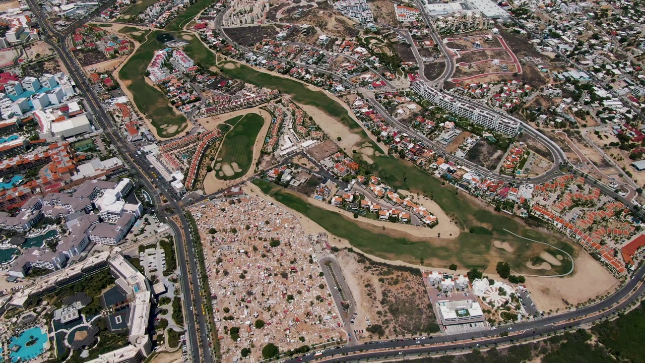 Second-frame aerial over San Jose del Cabo with the golf course centered against tan undeveloped ground, a divided road, nearby housing, parking areas, and no visible people.