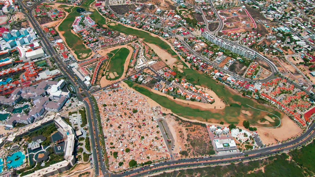 High aerial frame over San Jose del Cabo showing a green golf course between curved roads, red-roofed homes, larger buildings, blue pools, sandy open land, and no visible people.