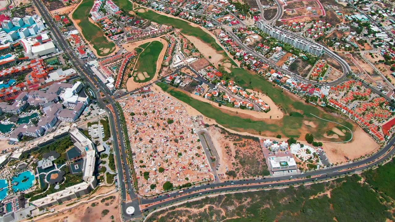 Overhead San Jose del Cabo view showing golf greens and sand areas beside a curved road, dense housing with red roofs, a large open lot of small structures, and no visible people.