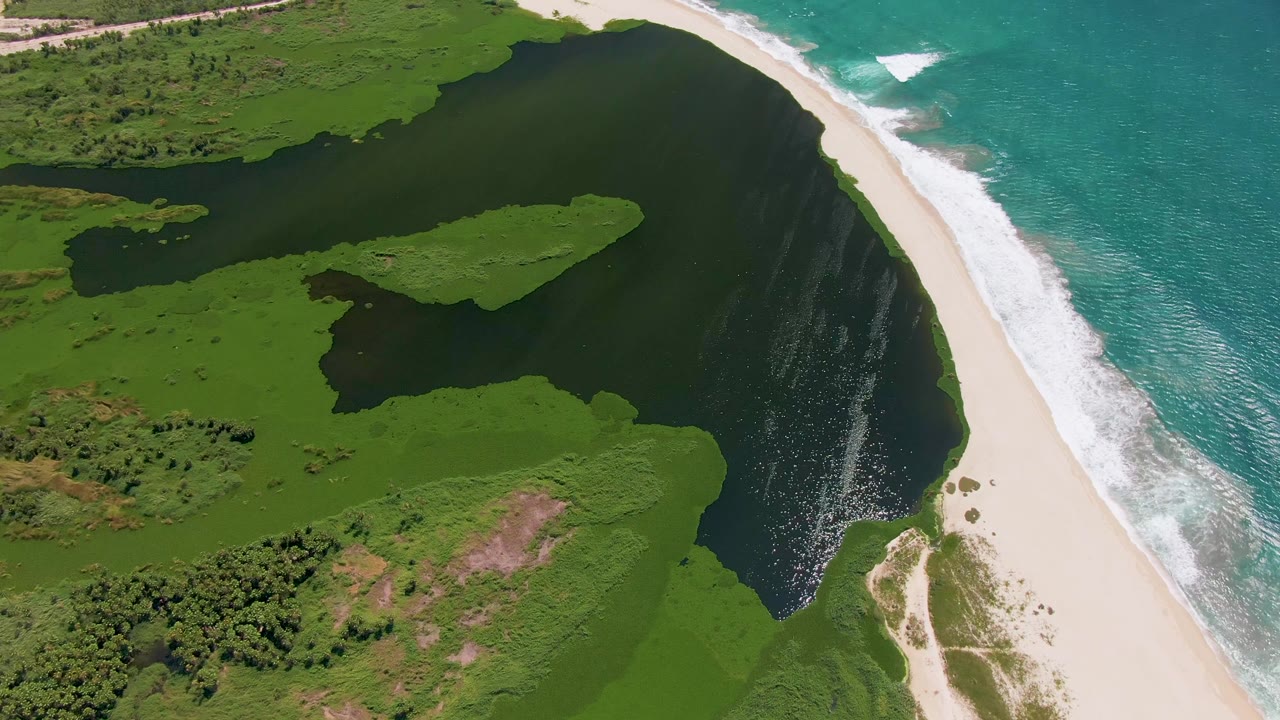 At 0:02, the aerial frame shows the San Jose del Cabo inlet widening inland, with a green island-like patch in dark water, white sand, surf, and no visible people.