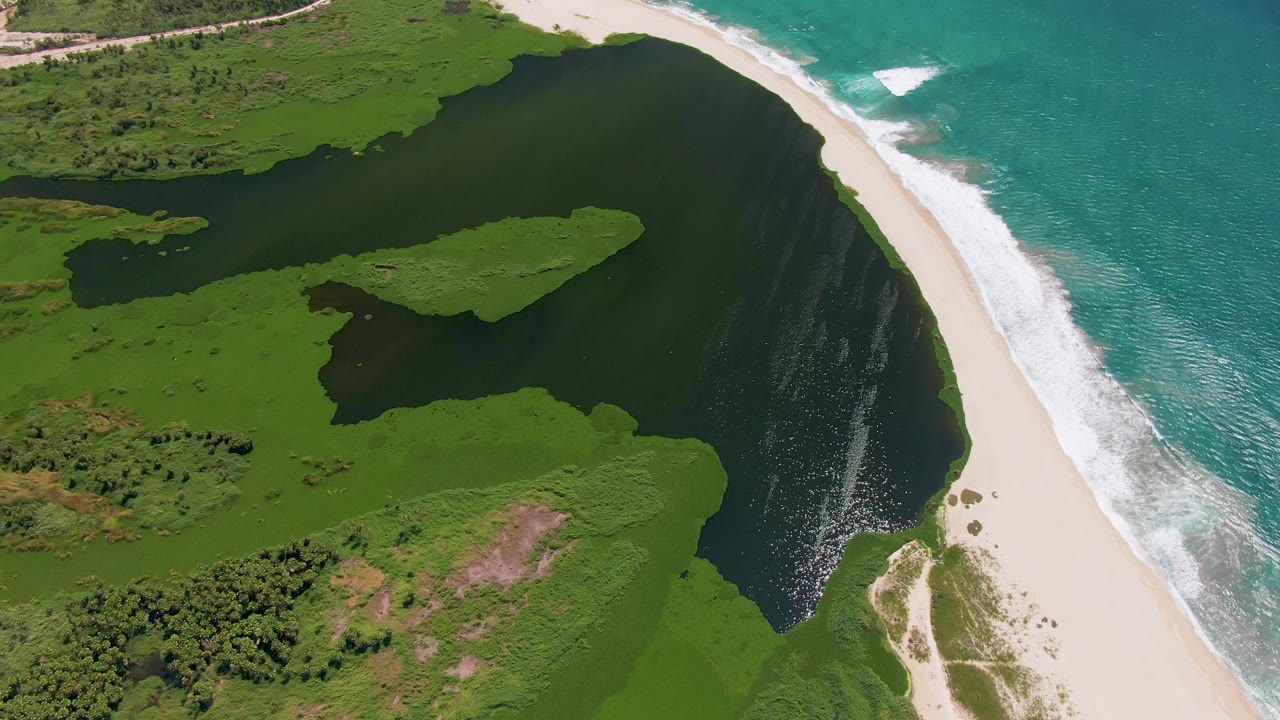 At 0:03, the San Jose del Cabo coastline remains overhead, with darker lagoon water on the left, a bright sandy shore to the right, ocean foam, and no visible people.