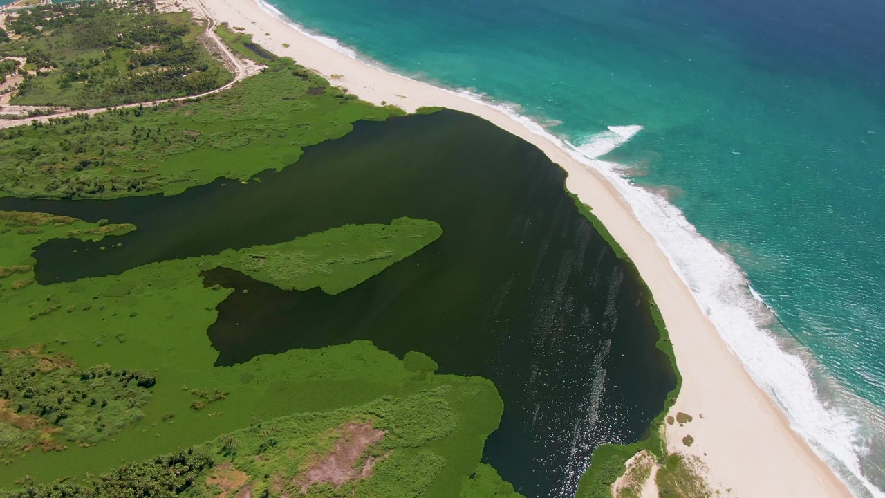 Aerial view in San Jose Del Cabo with a broad lagoon curve, green vegetation islands, pale sand, and white wave foam on the ocean side, with no visible people.