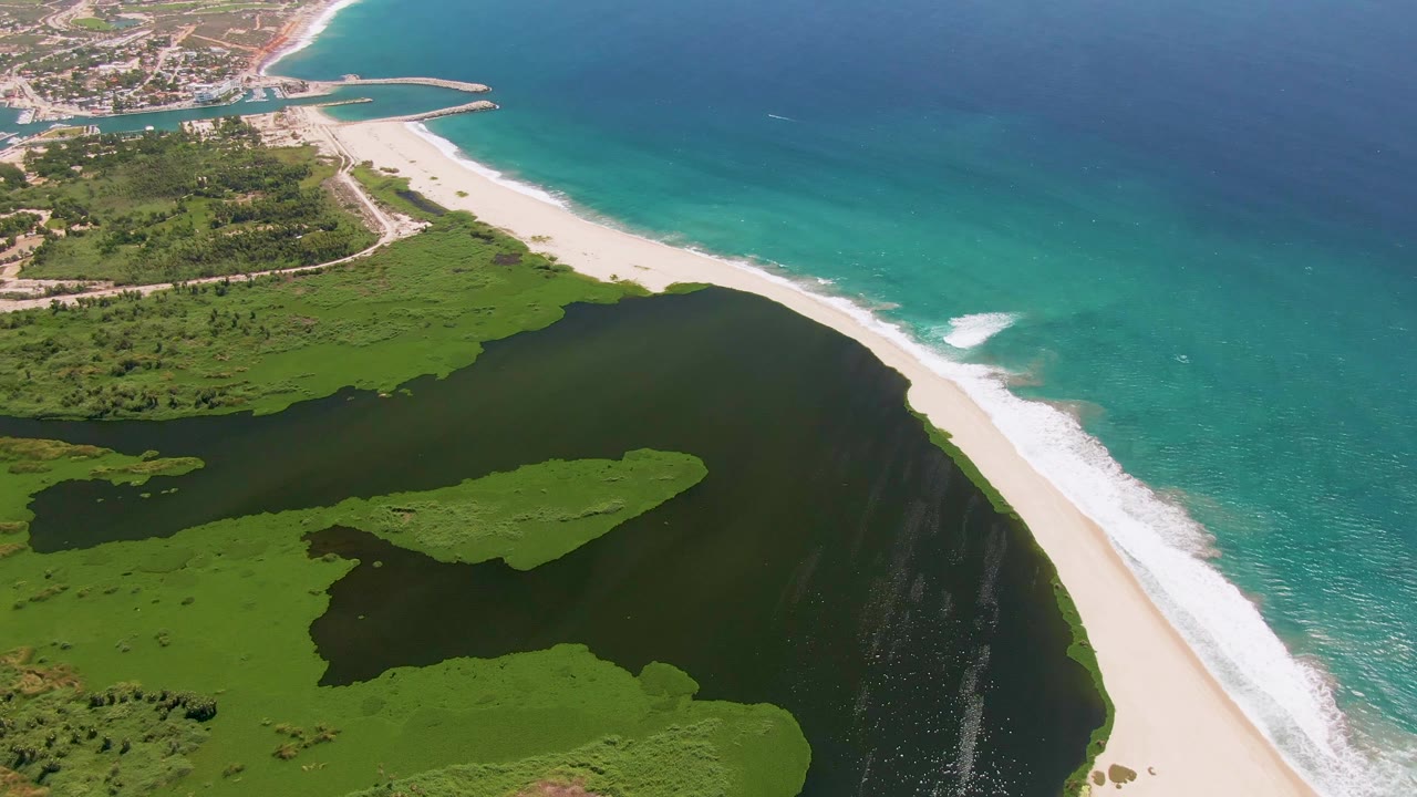 Aerial frame over San Jose Del Cabo with the lagoon filling the lower frame, a narrow sandy beach at right, and a bright surf patch along the turquoise water, with no visible people.