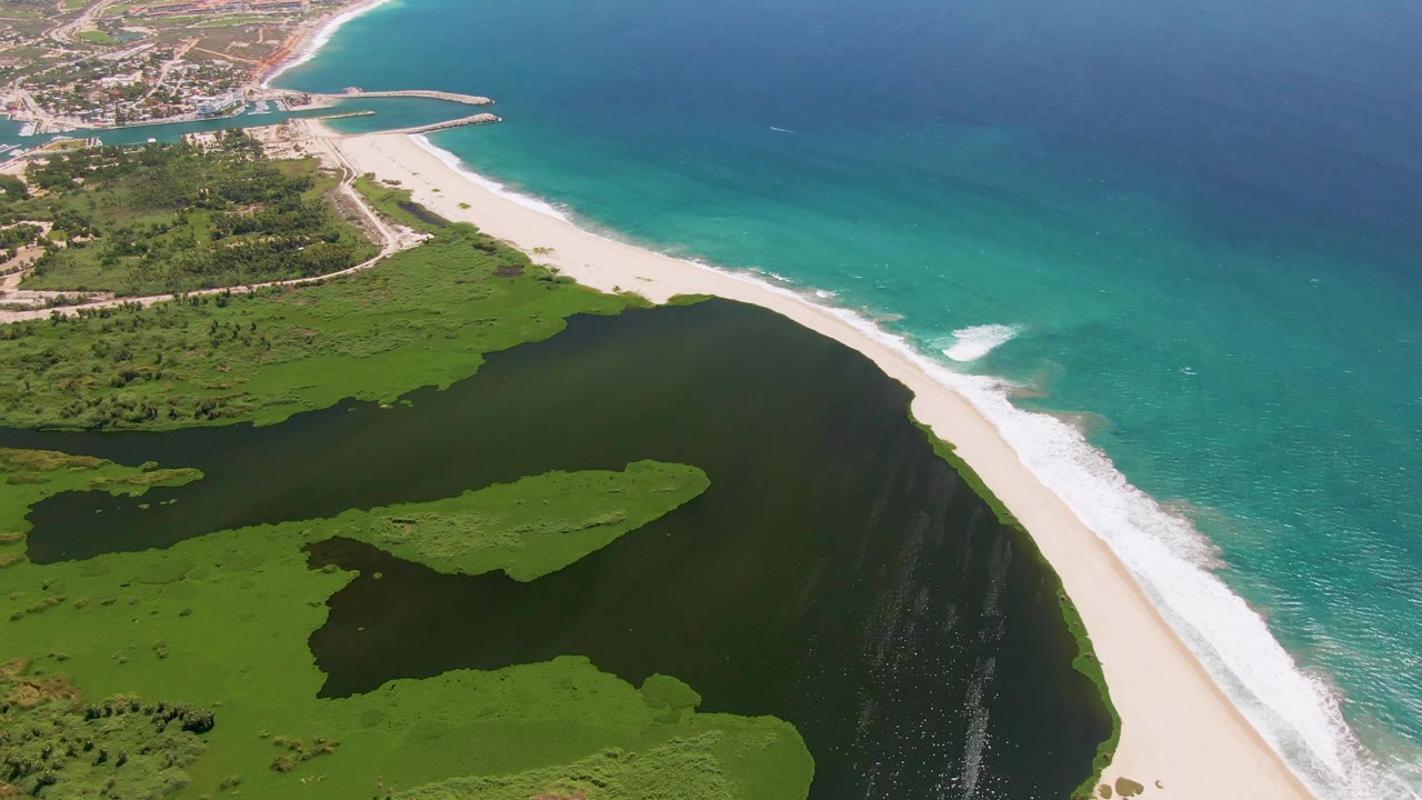 Aerial frame over San Jose Del Cabo showing green wetland islands inside the lagoon, the beach curving toward a distant shoreline, and deep blue ocean beyond, with no visible people.