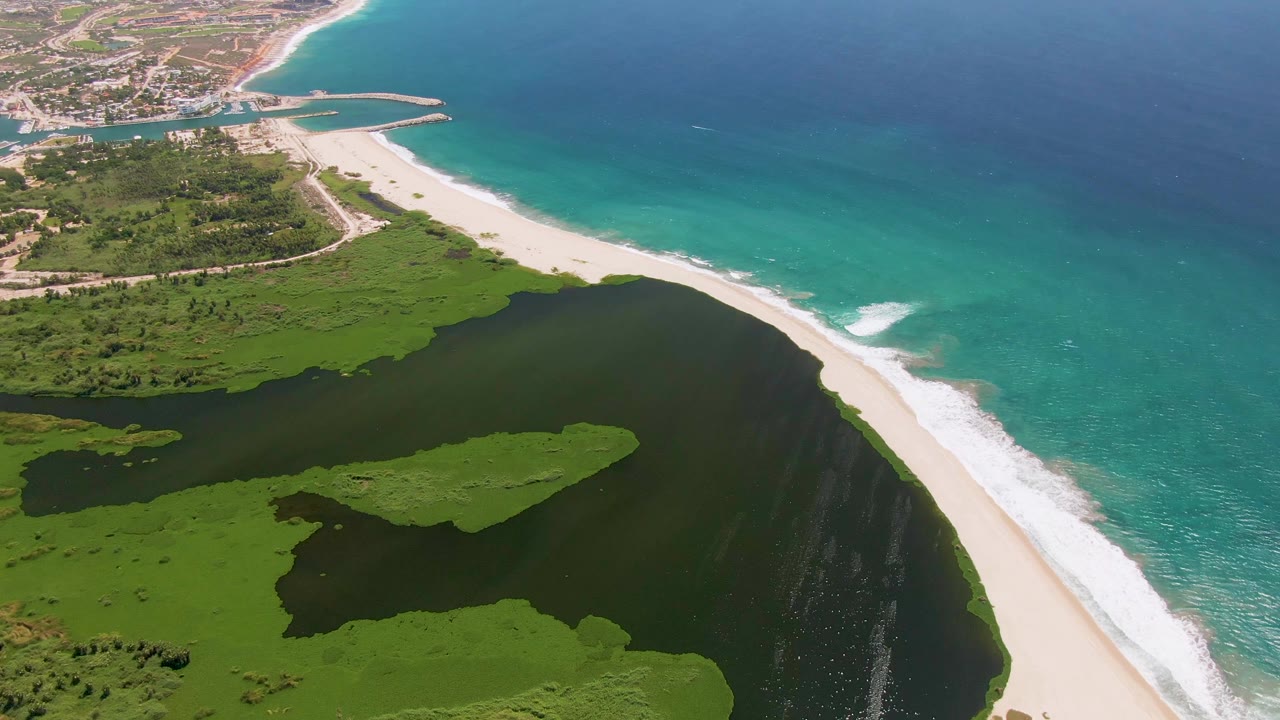 Aerial frame over San Jose Del Cabo with more wetland foreground visible, the lagoon leading diagonally to the sand bar and surf, and open ocean at right, with no visible people.
