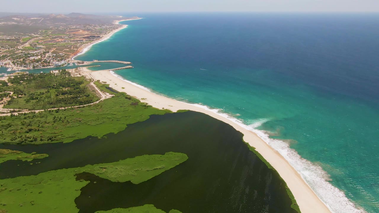 Opening aerial frame over San Jose del Cabo, Mexico showing green wetlands in the foreground, a sandy beach, turquoise ocean water, distant coastal settlement, and no visible people.