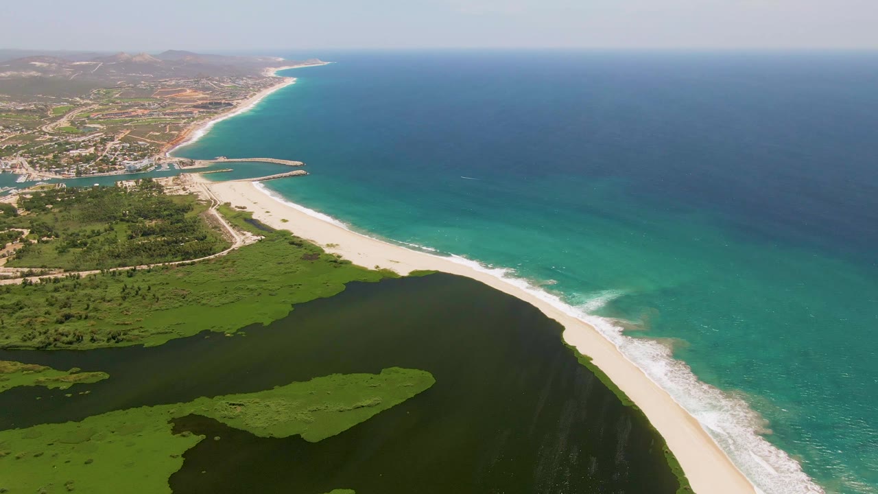San Jose del Cabo aerial frame with a pale sand strip dividing dark wetland water from turquoise ocean, white wave foam on the shore, inland roads and buildings in the distance, and no visible people.