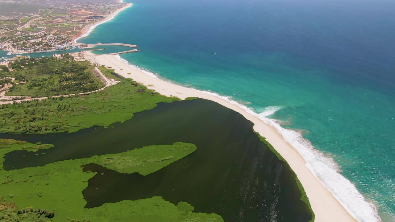 Drone view in San Jose del Cabo with white surf tracking along the sandbar, lagoon water in the foreground, and coastal buildings far inland, with no visible people.