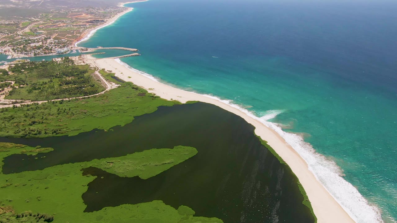 High aerial view of the San Jose del Cabo coast showing green lagoon islands, a bright beach edge, blue ocean water, and no visible people.