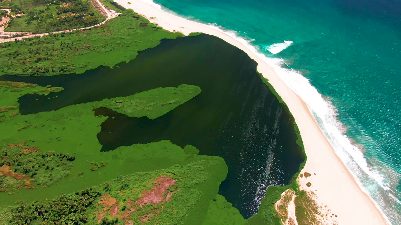 Aerial San Jose Del Cabo coastline with the lagoon curving beside the sand beach, white surf breaking on turquoise water, a small white boat wake offshore, and no visible people.