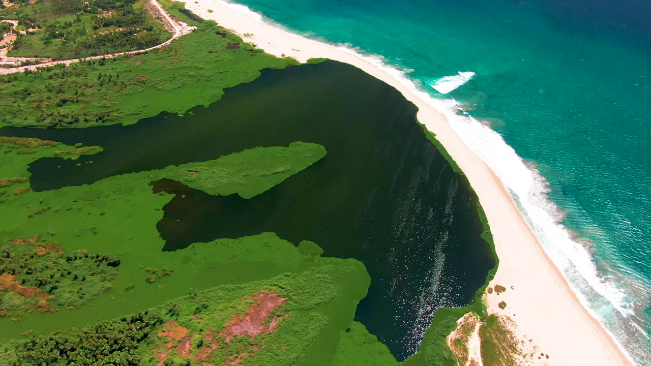 Drone view over San Jose Del Cabo with dark lagoon water in the middle, green marsh islands, bright sand, blue-green ocean, white surf, and no visible people.