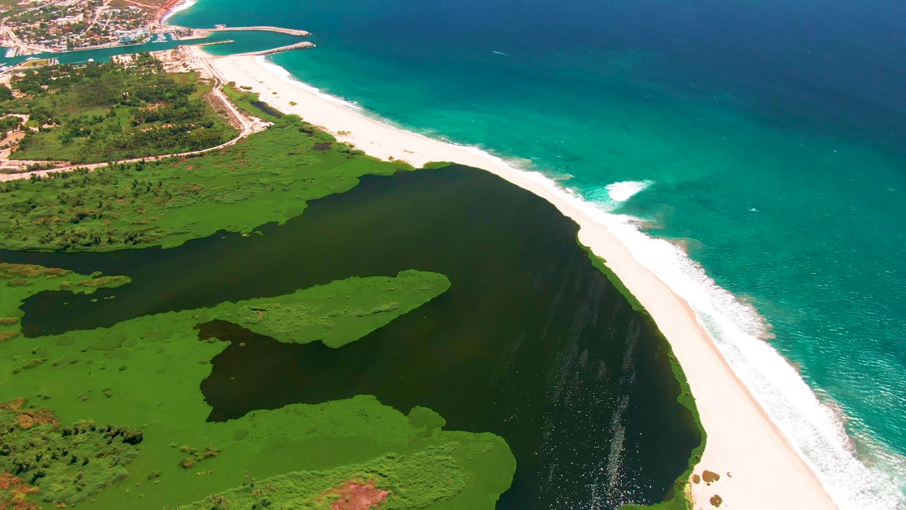Aerial frame over San Jose del Cabo showing dark lagoon water, green vegetation, a long white sand beach, turquoise surf, deeper blue ocean, and no visible people.