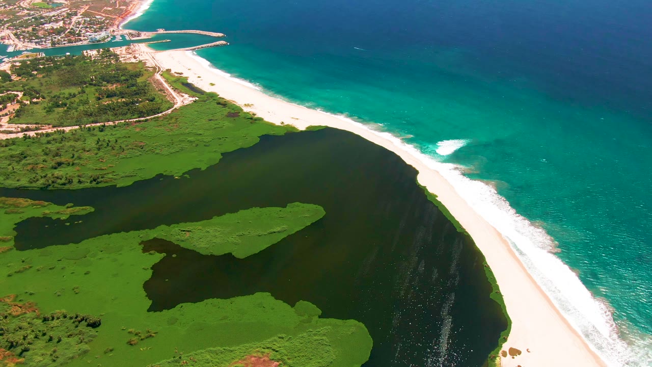 San Jose del Cabo aerial frame with dark lagoon water in the foreground, bright beach and foamy waves through the center, distant coastal buildings, and no visible people.