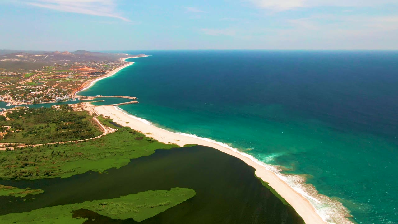 Aerial frame over San Jose del Cabo showing a dark green lagoon in the foreground, a pale sand beach curving beside turquoise ocean, distant buildings, low hills, and no visible people.