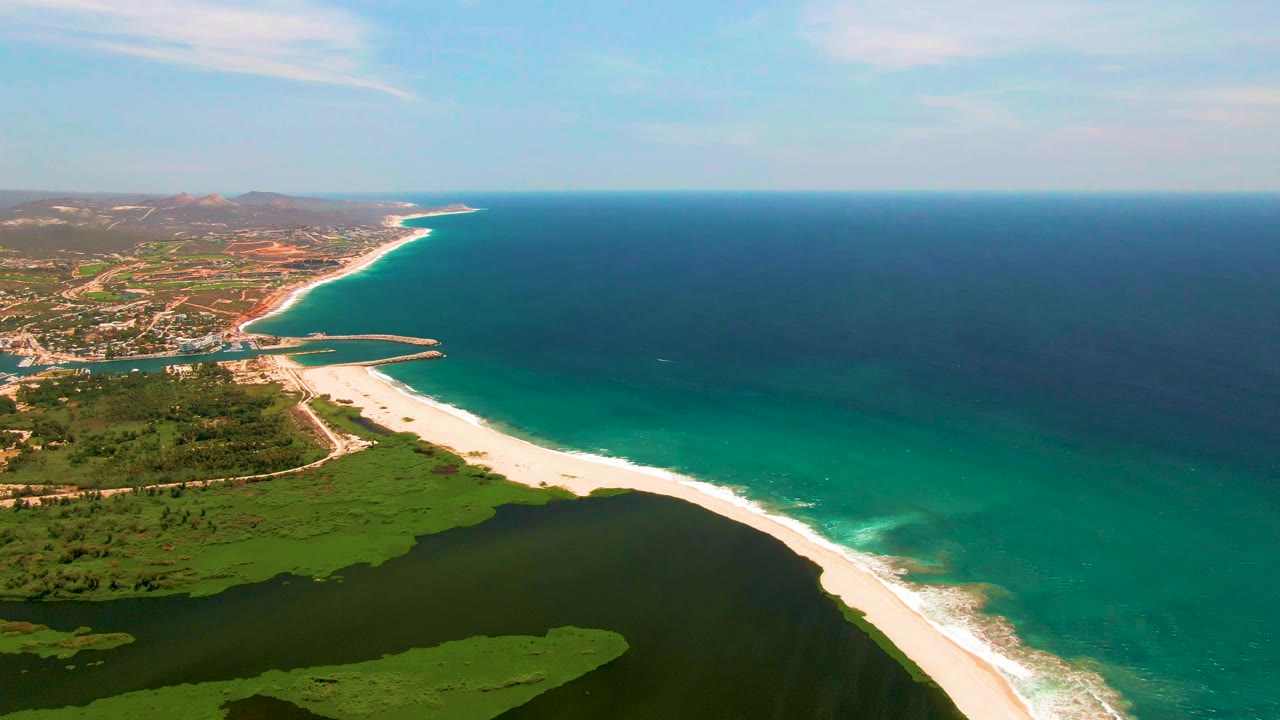 San Jose del Cabo aerial frame showing lagoon water and marshy green areas below, a long pale beach, turquoise shallows near shore, darker ocean beyond, and no visible people.