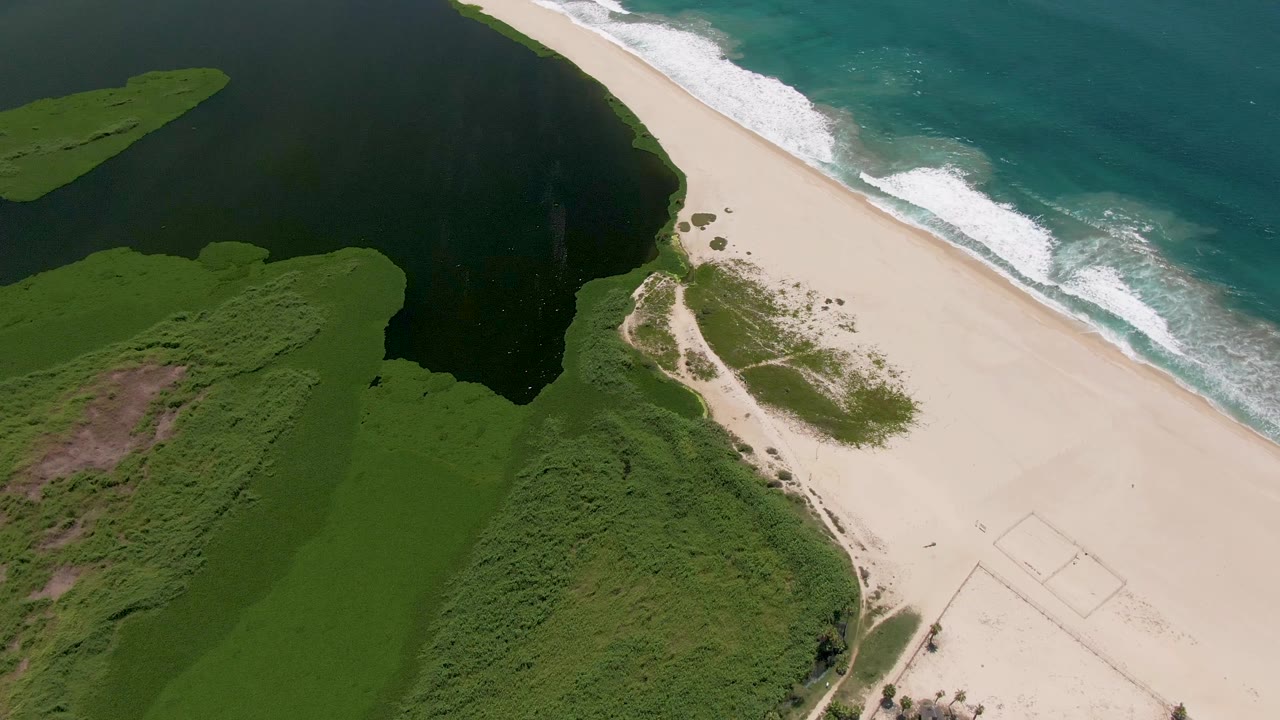 Aerial view in San Jose del Cabo with the lagoon filling the left side, a broad sand strip through the center, white foam along the ocean edge, and no visible people.