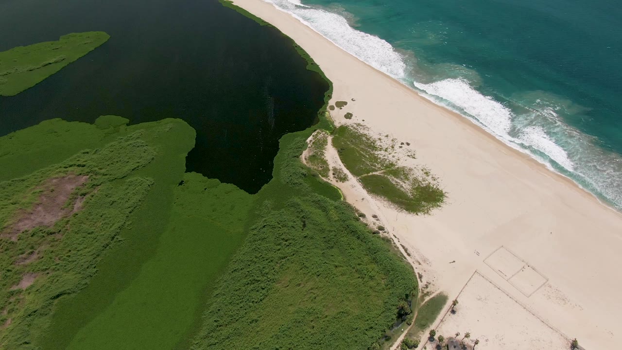 Aerial San Jose del Cabo coastline frame showing green vegetation curving around the lagoon, empty sand beside it, turquoise ocean water, and no visible people.
