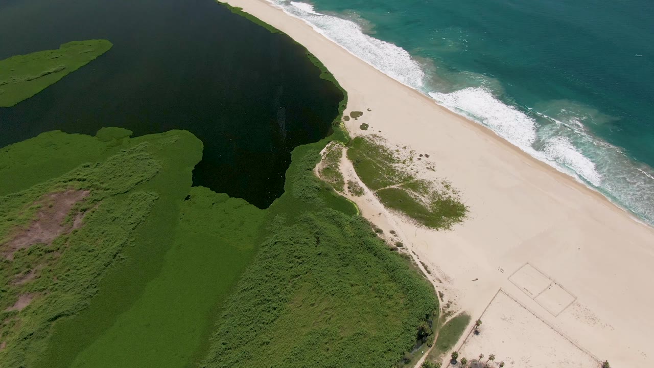 Aerial coastline with lagoon, vegetation, sand, and waves near the shore.