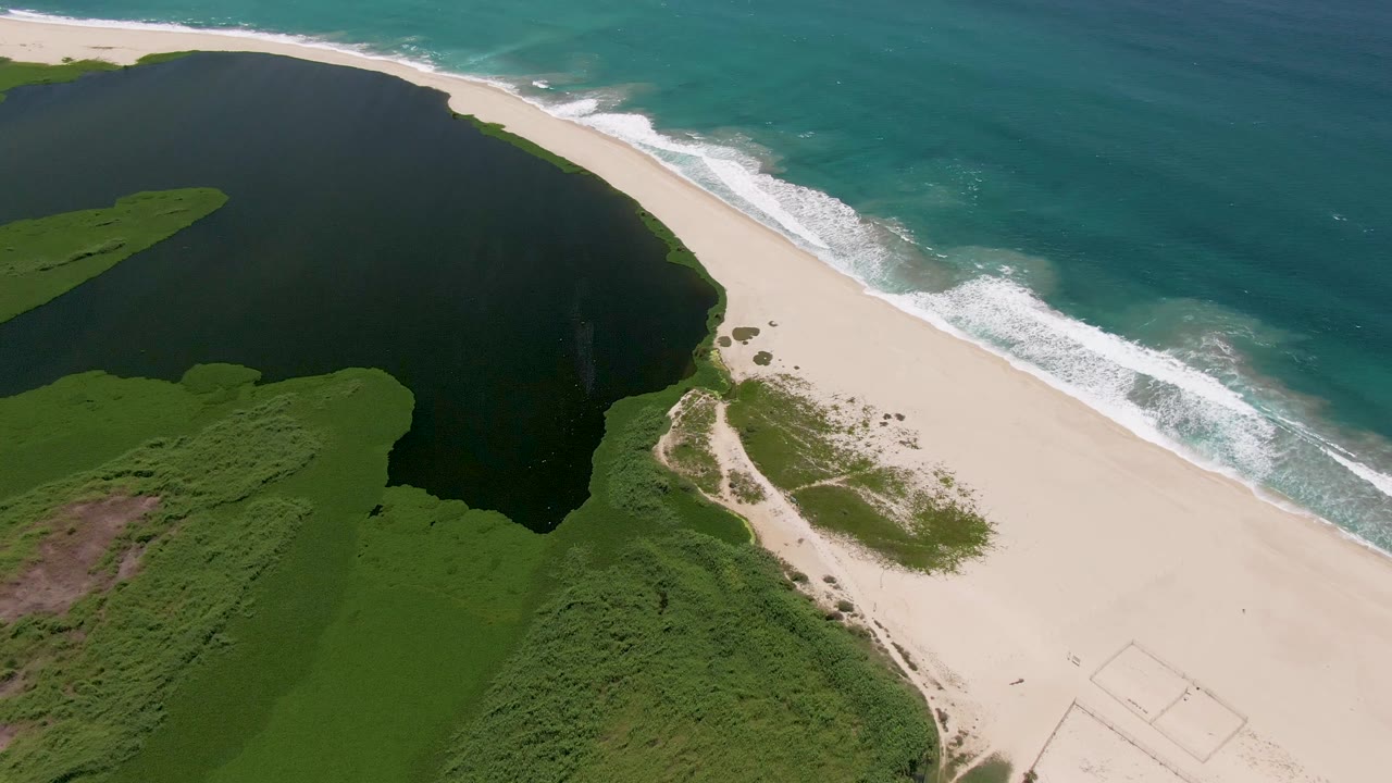 High aerial view in San Jose del Cabo where the dark lagoon curves beside the sand and the blue-green ocean sits beyond a line of white surf, with no visible people.