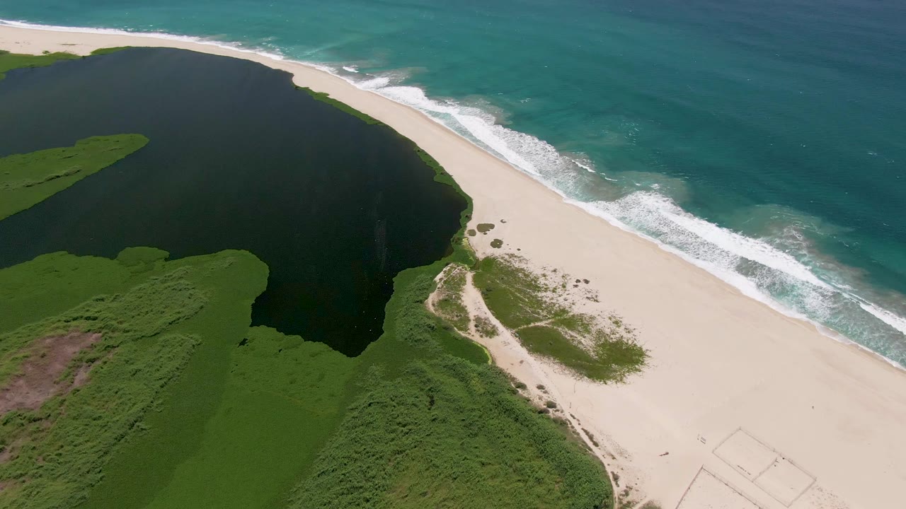 Aerial shoreline with dark lagoon, pale sand, waves, and dense green vegetation.