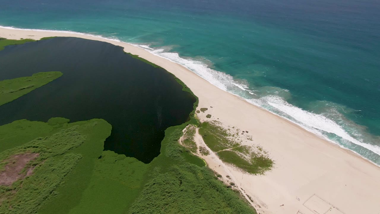 Aerial view in San Jose del Cabo showing a dark lagoon beside green vegetation, a pale sandy beach, turquoise ocean water, and no visible people at the start of the clip.