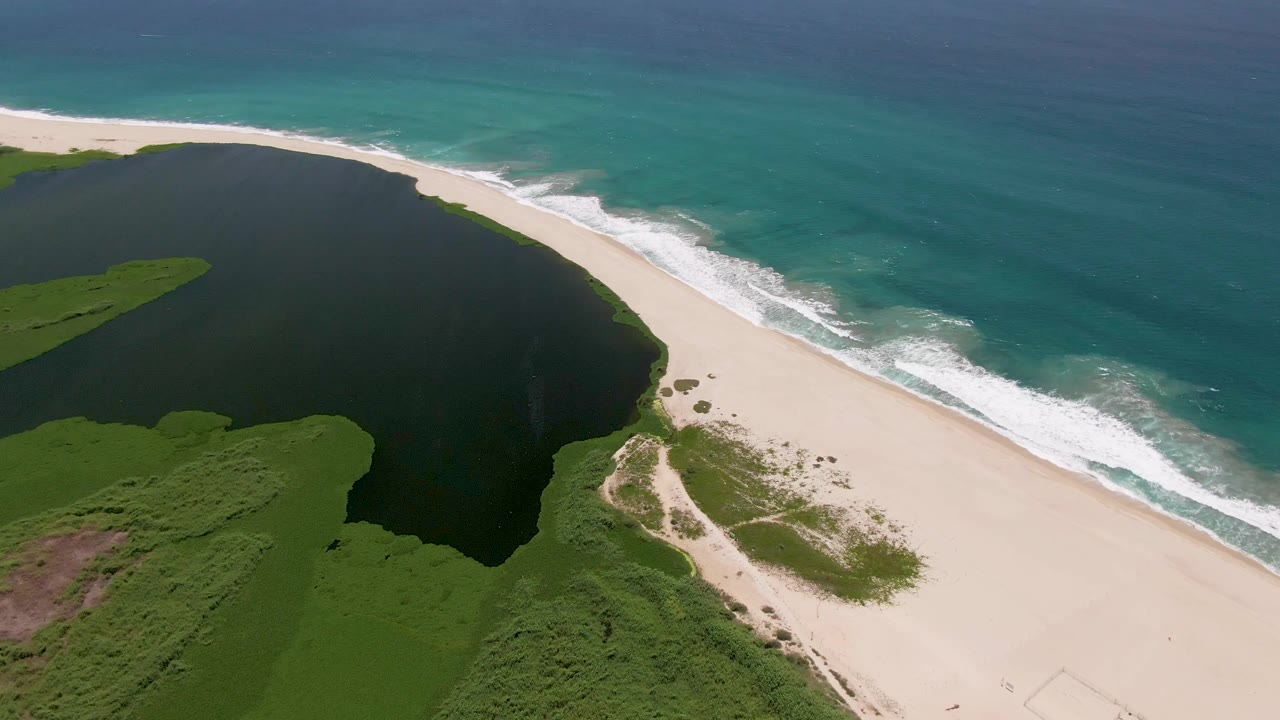 Aerial frame in San Jose del Cabo with the lagoon occupying the left side, surf reaching the sandy beach on the right, and no visible people.