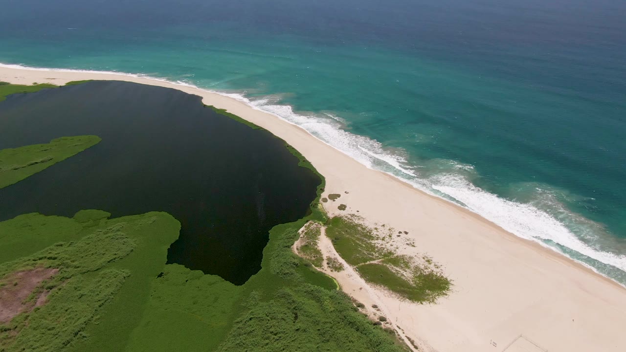 San Jose del Cabo aerial still showing green vegetation wrapping the lagoon, white sand curving along the shore, turquoise ocean beyond, and no visible people.