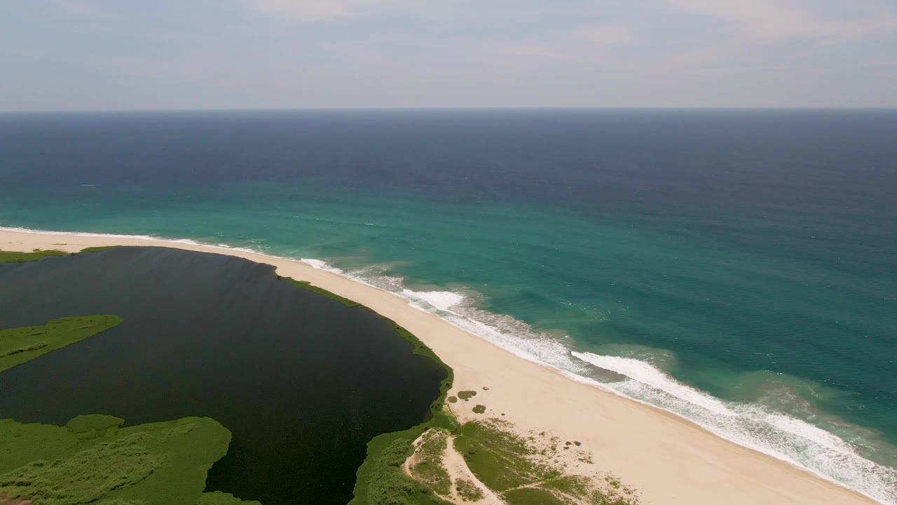 Aerial frame from San Jose Del Cabo showing a dark lagoon on the left, pale sandy beach, turquoise ocean surf, and no visible people.
