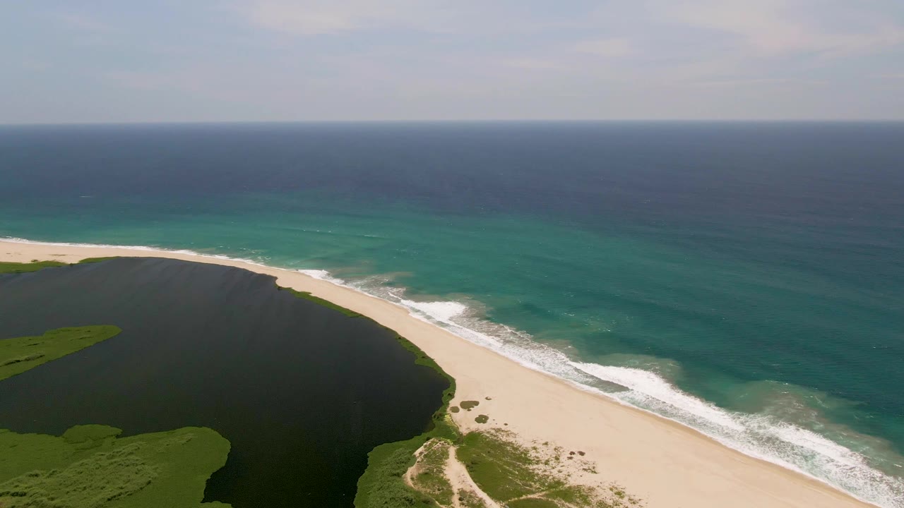 Aerial view in San Jose Del Cabo with the lagoon curving into the frame, white surf moving along the sand, open ocean beyond, and no visible people.