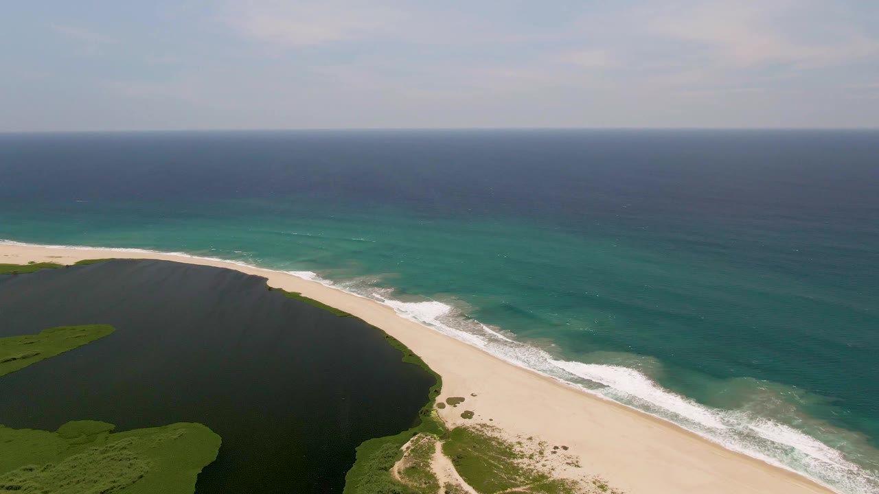 San Jose Del Cabo aerial frame showing a wide sand strip between the dark lagoon and green-blue ocean, with waves breaking near shore and no visible people.