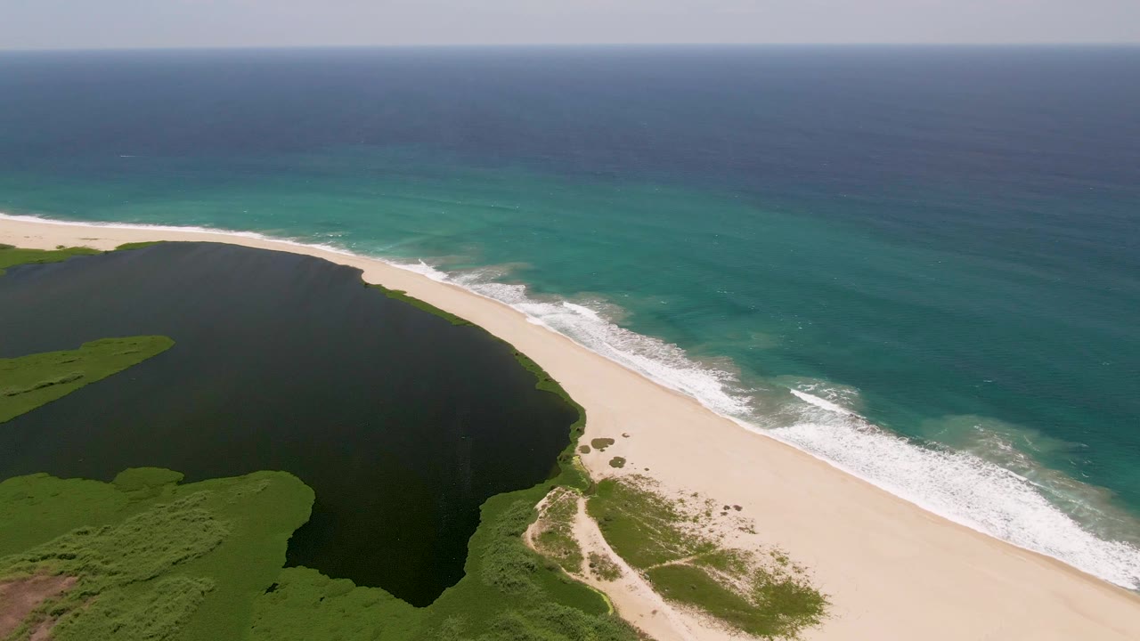 Aerial view in San Jose Del Cabo showing a dark coastal lagoon, pale sand beach, turquoise ocean, white surf, green vegetation, and no visible people at the start of the clip.