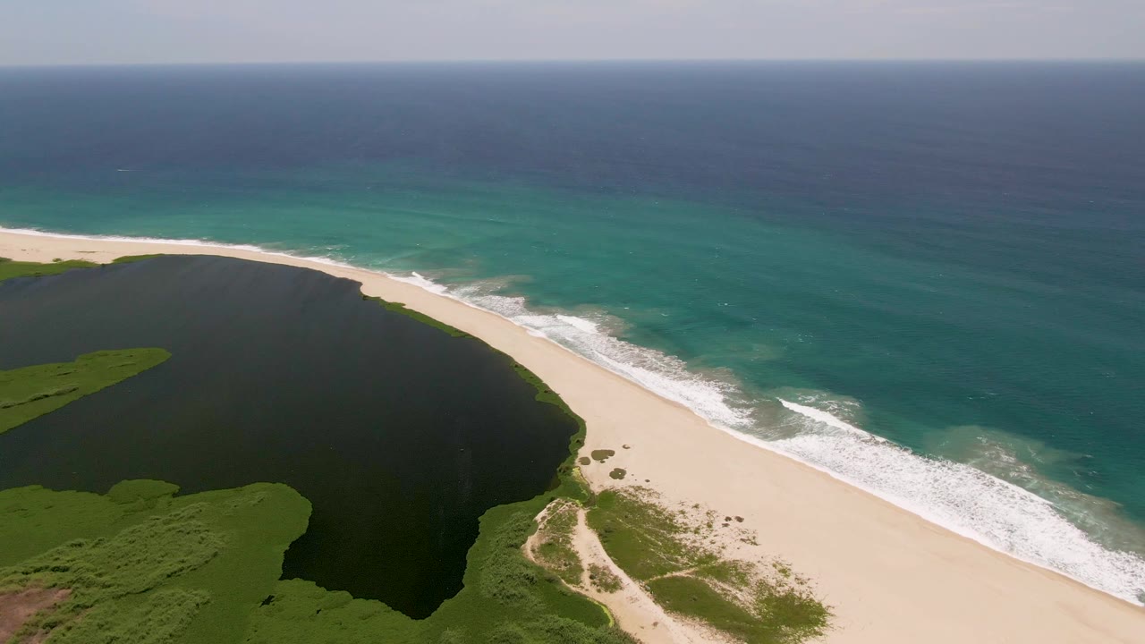 Aerial frame in San Jose Del Cabo with the curved sandy shore separating the dark lagoon from the turquoise ocean, while white waves advance along the beach and no visible people appear.